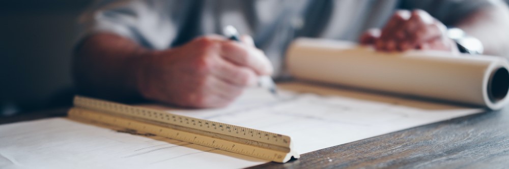 A draft table with a man's hand holding a pencil over a piece of draft paper. A metal ruler is in the foreground. Photo by Daniel McCullough on Unsplash.